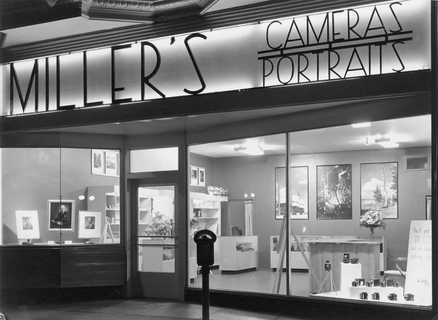 The original Miller's storefront at night, its lit sign reading Miller's Cameras Portraits, portraits displayed in the window