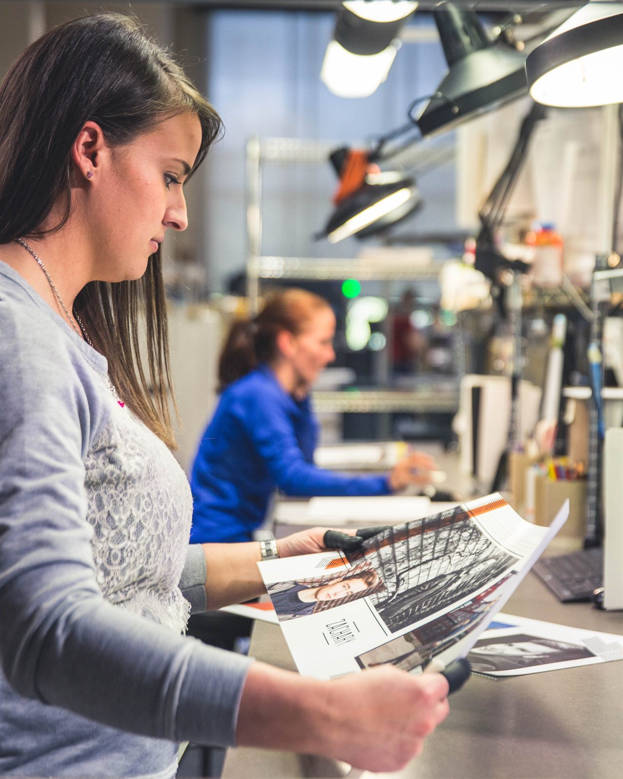 A Miller's employee checking a freshly printed sheet under a task lamp at the inspection bench, a colleague working further down the line